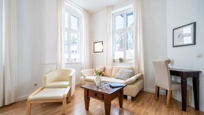 Une pièce avec un plancher en bois, un canapé, une chaise, une petite table sur laquelle se trouvent un vase et des verres, et un tableau encadré accroché au mur.