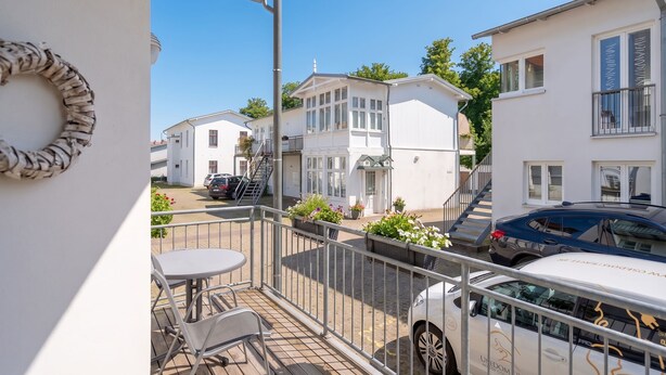 Un balcon avec une table et des chaises blanches, une voiture blanche garée devant, et un bâtiment blanc avec un balcon en arrière-plan.