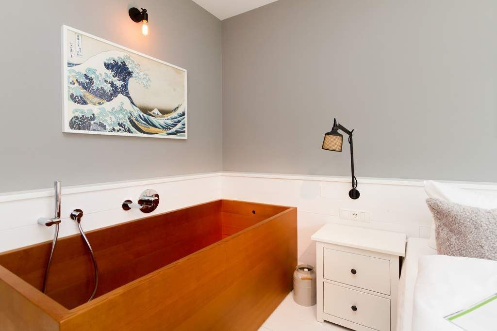 Un bagno con una vasca da bagno in legno, un comodino bianco, un tavolino da notte e un'opera d'arte appesa al muro.