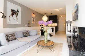 A modern living room with a light grey sofa, a round wooden coffee table with a vase of flowers, a chandelier, and a kitchen in the background. - Villa Strandgrün, App. 05 (Heringsdorf)