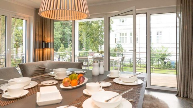A modern dining area with a table set for breakfast, large windows, and a view of greenery outside. - Villa Strandgrün, App. 06 (Heringsdorf)