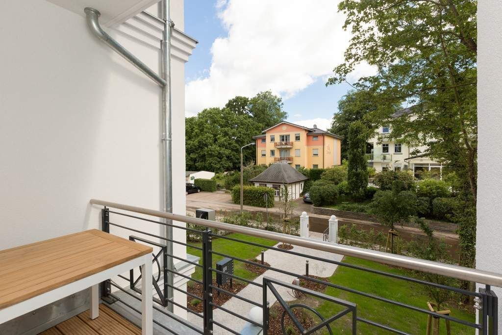 Un balcon doté d’une table en bois, d’une rambarde en métal et offrant une vue sur un quartier résidentiel avec des maisons et de la verdure.