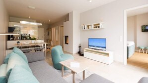 A modern living room with a sofa, a coffee table, and a TV. There is a kitchen in the background with a dining area. - Villa Strandgrün, App. 02 (Heringsdorf)