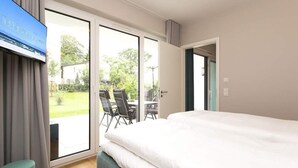 A hotel room with a bed, a television, a sliding glass door to an outdoor area with a table and chairs, and a view of greenery. - Villa Strandgrün, App. 02 (Heringsdorf)