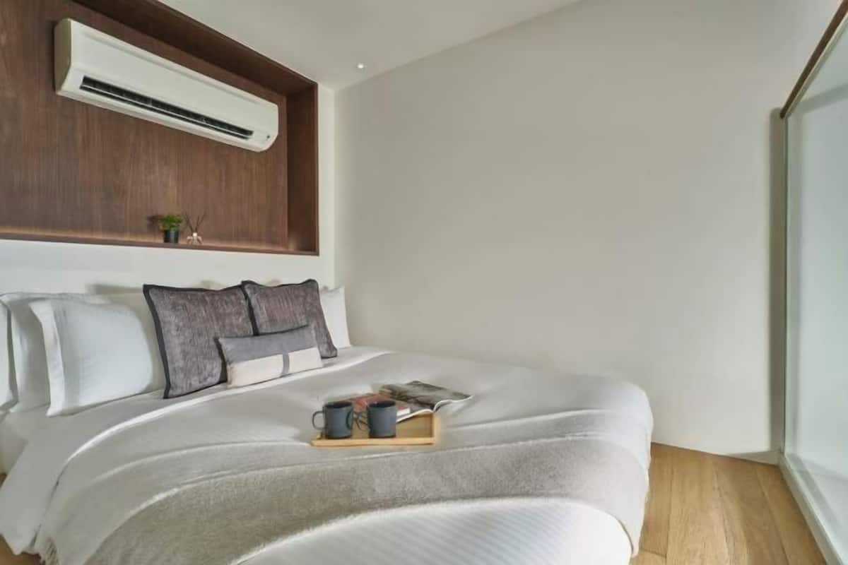 Un dormitorio con una cama, almohadas, una mesita de noche con un libro y dos tazas, un equipo de aire acondicionado y una ventana.