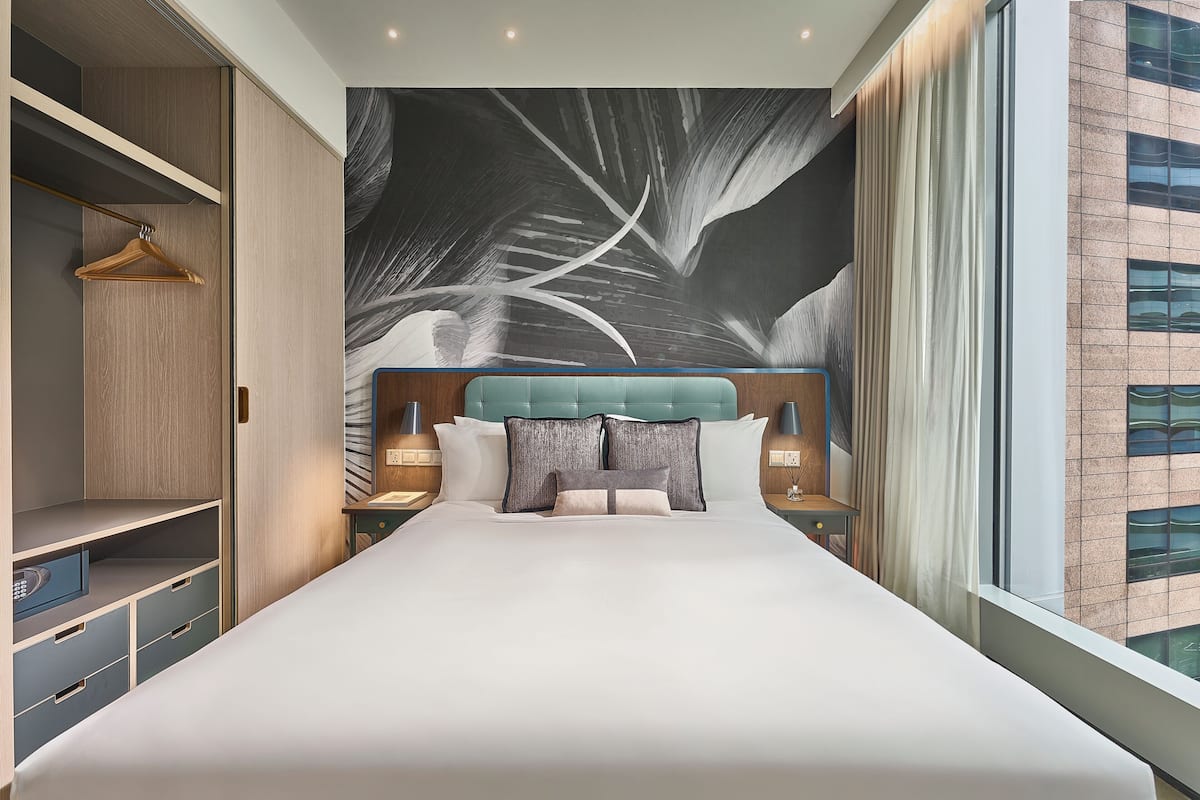Una habitación de hotel moderna con una cama grande, un armario, un cabecero con un panel tapizado en azul claro y un ventanal con vistas a un edificio.