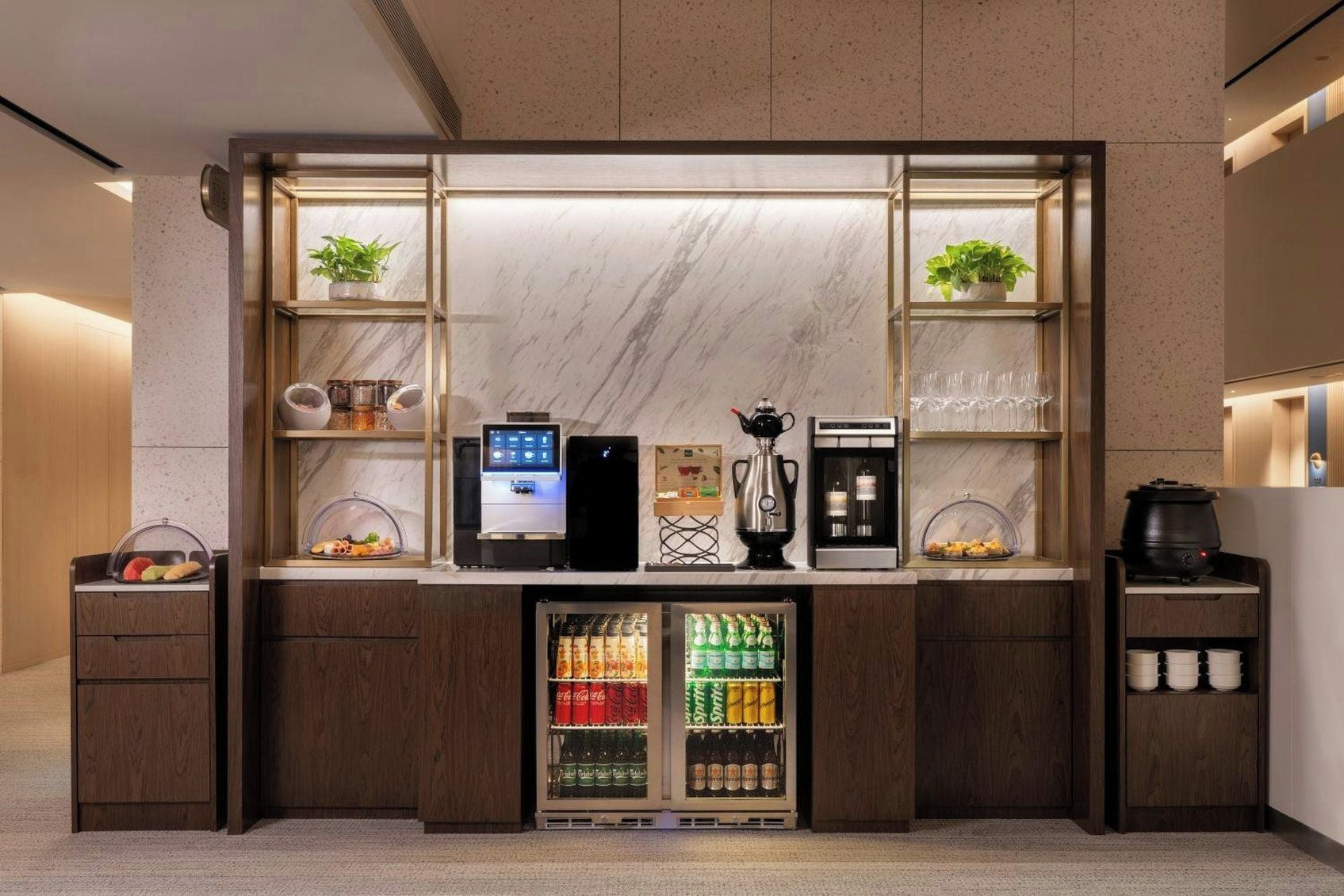 A self-service beverage station with a coffee machine, tea pot, and refrigerator stocked with beverages.
