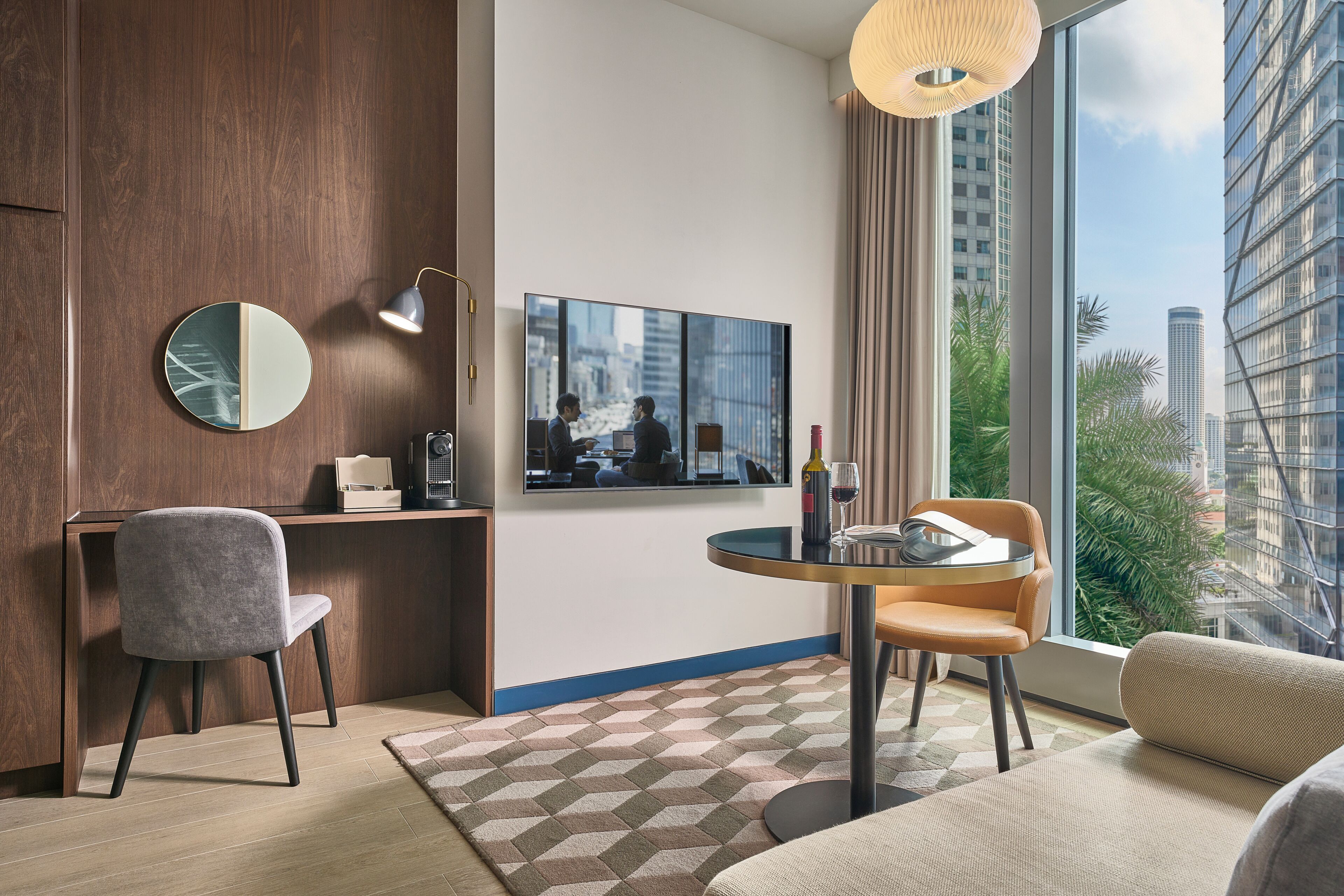 A modern hotel room with a large window, a round table, a sofa, a chair, a desk, a mirror, and a wall-mounted TV.