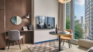 A modern hotel room with a large window, a round table, a sofa, a chair, a desk, a mirror, and a wall-mounted TV.
