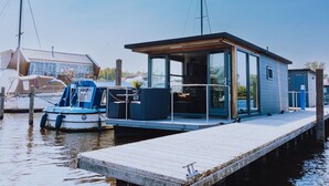 Ein modernes Hausboot liegt an einem Steg mit freiem Blick auf das Wasser und einer kleinen Deckfläche.