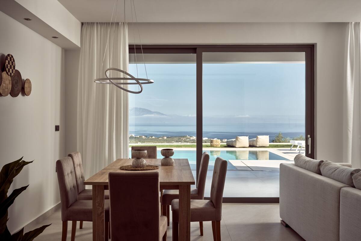 A modern dining area with a wooden table and chairs, a large window with a view of the sea, and a sofa in the foreground.