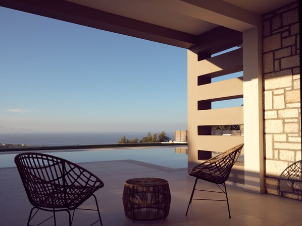 A modern outdoor seating area with two wicker chairs and a round table, overlooking a pool and the sea.