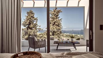 A bedroom with a large window overlooking a balcony with a view of trees and the sea.