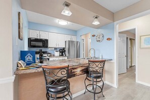 A kitchen with white cabinets, a stainless steel refrigerator, a microwave, and a coffee maker. There are two bar stools with a decorative backsplash.