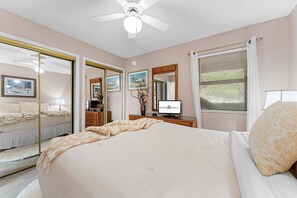 Een slaapkamer met een bed, een dressoir, een spiegel, een plafondventilator en een raam met jaloezieën.