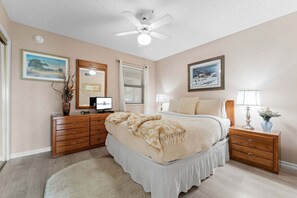 Ein Schlafzimmer mit einem Bett, einem Schminktisch, einem Nachttisch, einem Deckenventilator und zwei gerahmten Bildern an der Wand.