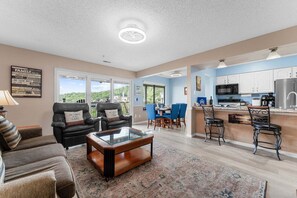 A spacious living room with a glass coffee table, a dining area with blue chairs, and a kitchen with white cabinets and stainless steel appliances.