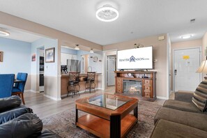 A living room with a glass coffee table, a fireplace, a television, and a kitchen area with cabinets and a refrigerator.