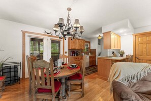 A dining area with a wooden table, chairs, and a chandelier. There is a kitchen in the background with wooden cabinets and a sink.