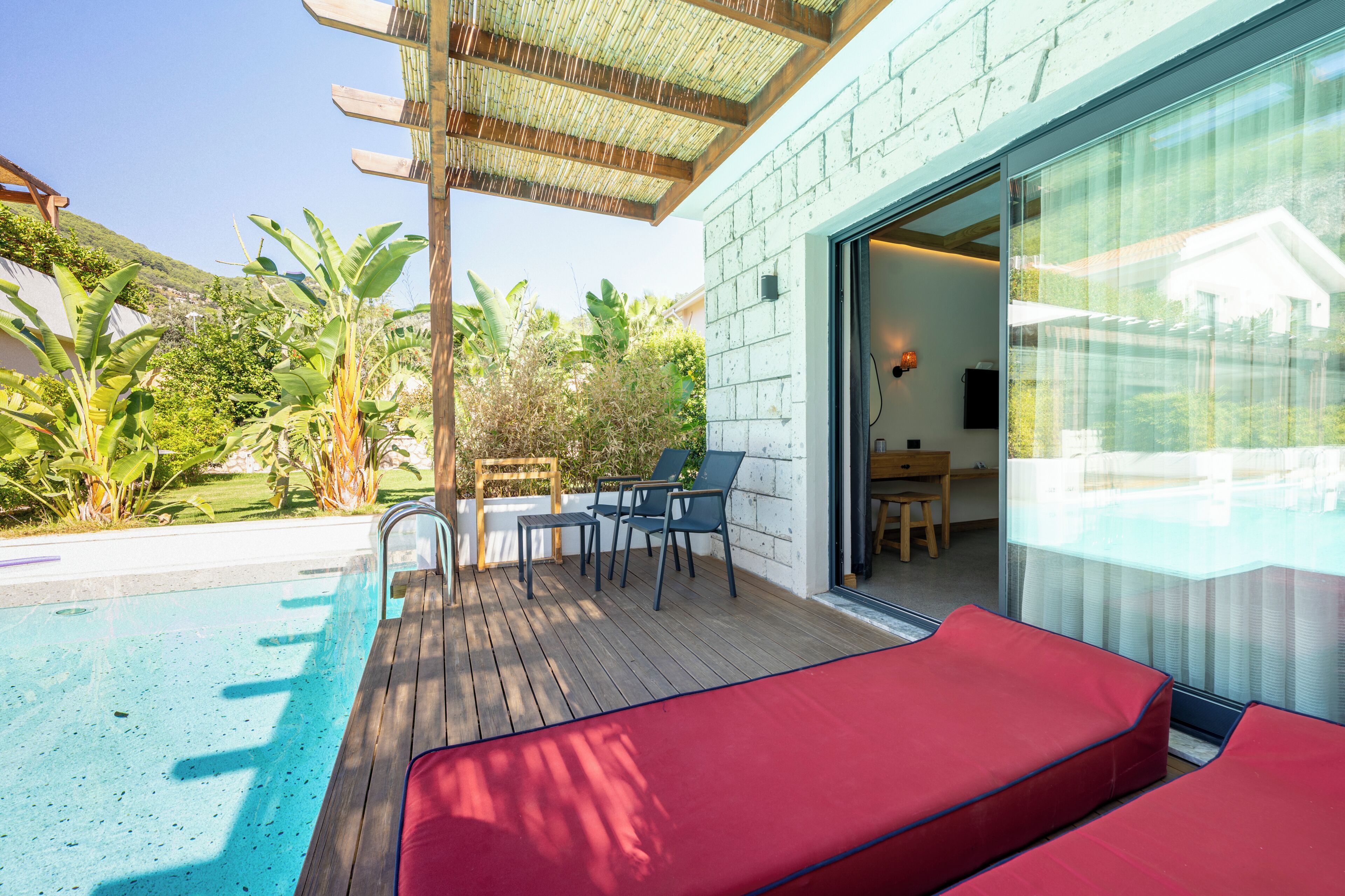 Un espace piscine doté d’une terrasse en bois, de chaises longues rouges et d’une porte vitrée donnant accès à une pièce.