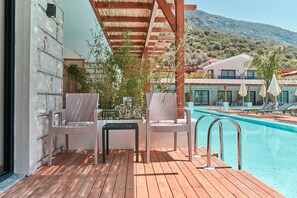 A wooden deck with wicker chairs and a poolside table, overlooking a swimming pool and a mountain backdrop. - The Pearl Ölüdeniz (Fethiye)