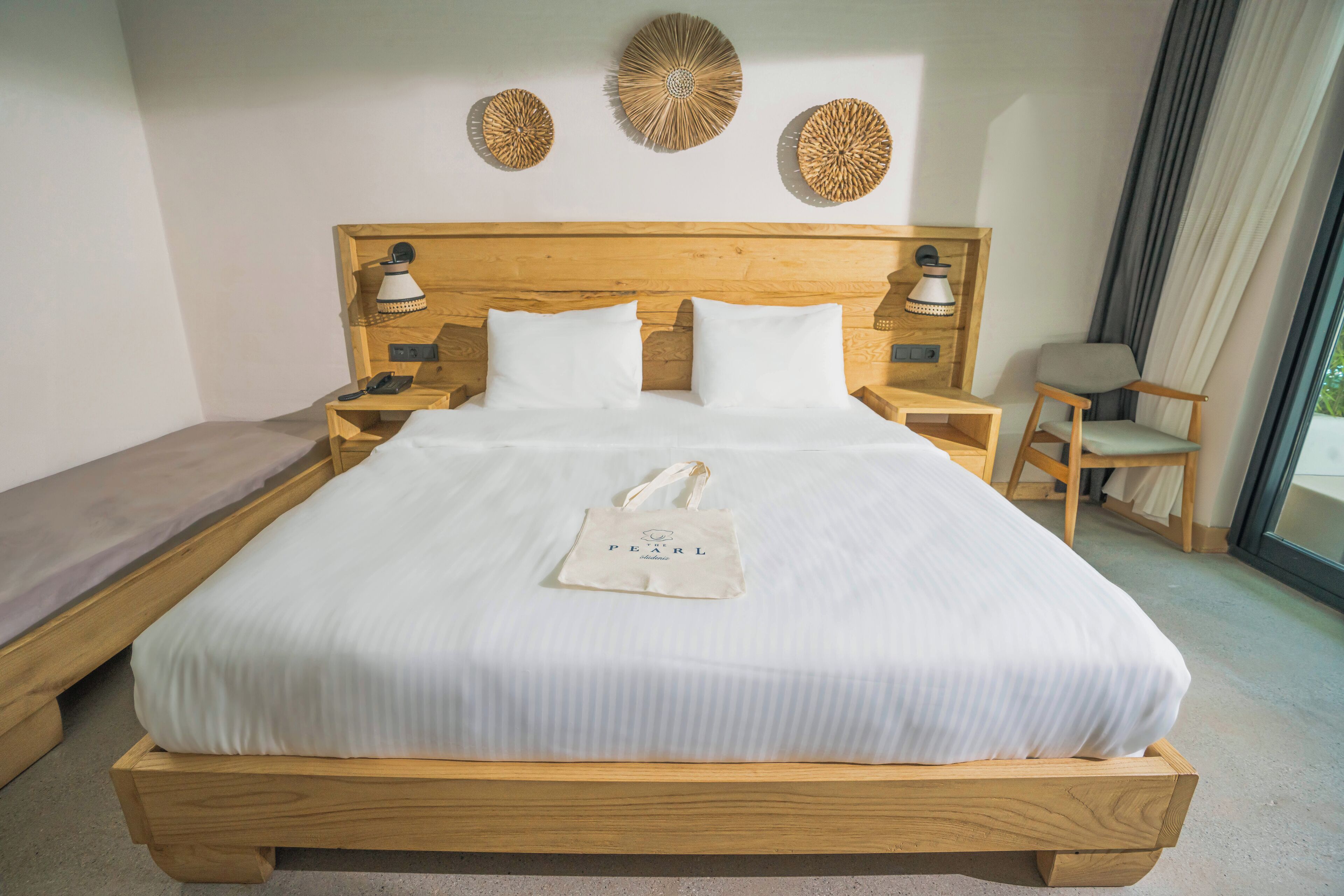 A neatly made bed with a white pillow and a white bag on it, wooden headboard, bedside tables with lamps, and a chair by the window.