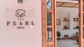 A sign for "The Pearl" hotel with a logo, a wooden door frame, and a view of the interior including shelves with plants and a wicker chair. - The Pearl Ölüdeniz (Fethiye)