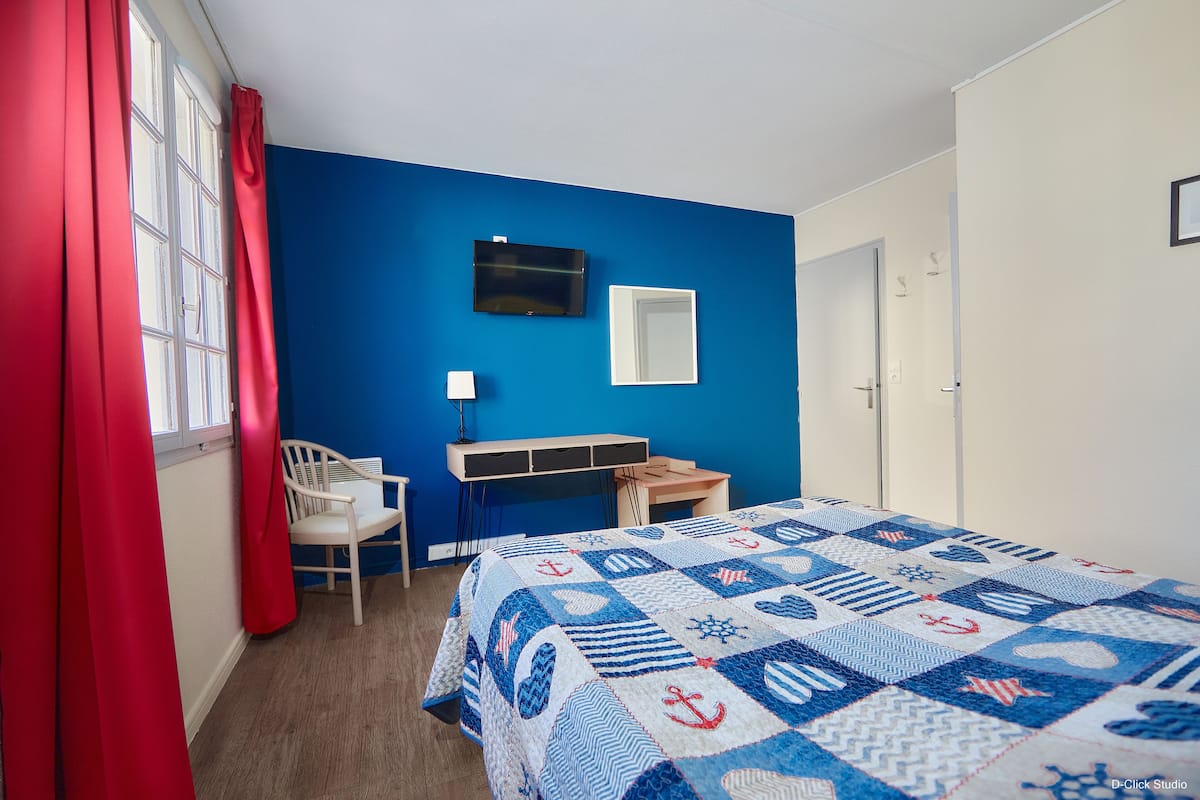 Un dormitorio con pared azul, cama con colcha de temática náutica, escritorio, silla y televisor en la pared.