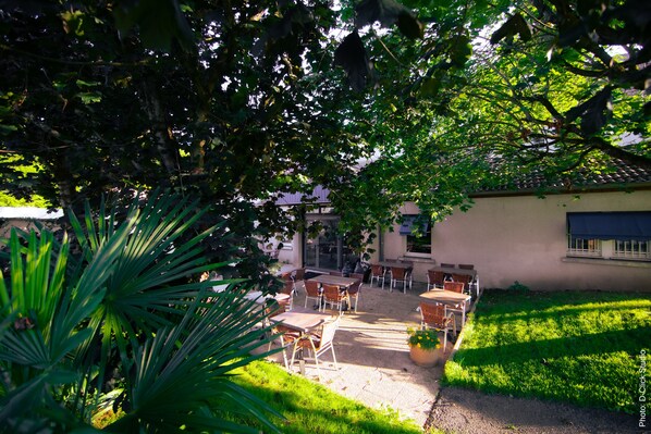An outdoor patio with tables and chairs, surrounded by greenery and a building in the background. - Les Tilleuls (Rochefort-sur-Nenon)