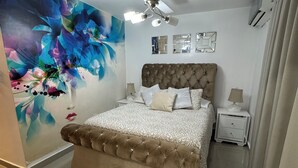 Une chambre avec un lit doté d’une tête de lit capitonnée, un ventilateur de plafond, une table de chevet avec une lampe et un mur orné d’une œuvre d’art florale abstraite.