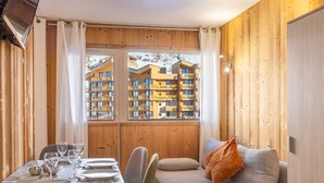 A cozy room with a dining table set for four, a sofa with cushions, and a window with a view of a building. - Charming 2-Bedroom Apartment in Val Thorens - Sleeps 4, Renovated, Ski-In/Ski-Out! (Val Thorens)