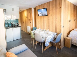 Quarto compacto com mesa de jantar posta para quatro pessoas, kitchenette, cama e televisão fixada na parede de madeira.