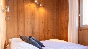 A bedroom with a wooden wall, a bed with white bedding, and a window with curtains. - Charming 2-Bedroom Apartment in Val Thorens - Sleeps 4, Renovated, Ski-In/Ski-Out! (Val Thorens)