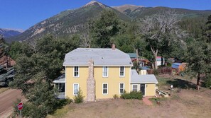 A two-story yellow house with a stone chimney, surrounded by trees and mountains in the background.