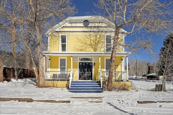 A two-story yellow house with a front porch, surrounded by snow and leafless trees.