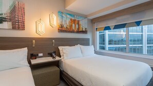 A hotel room with two beds, a nightstand with a phone, a wall-mounted artwork, and a window with a view of buildings.