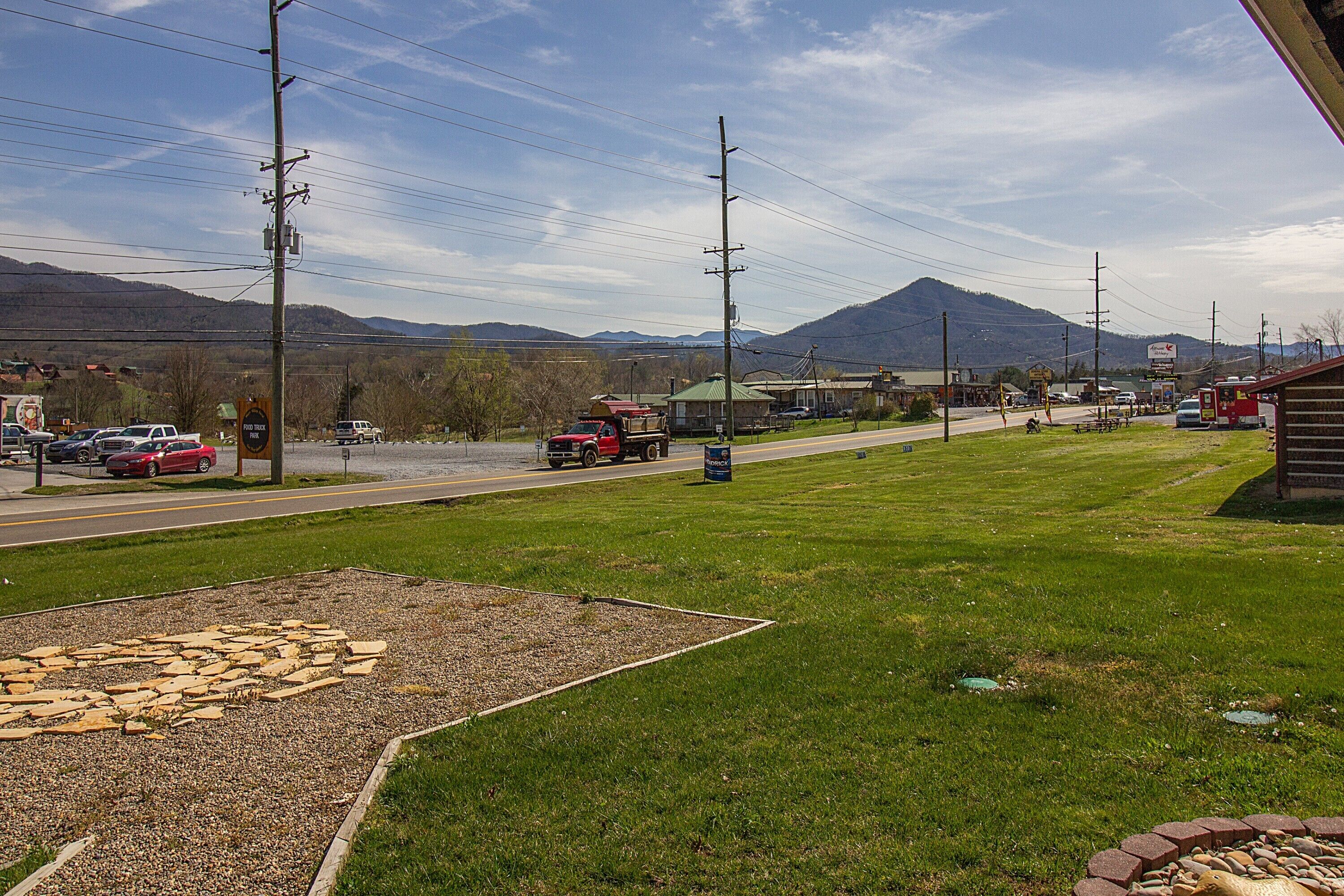 A grassy area with a stone pathway, a road with vehicles, and a mountain in the background.