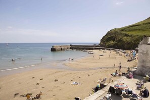 Una playa con un muelle de piedra que se adentra en el mar, gente tomando el sol y una colina cubierta de hierba al fondo.