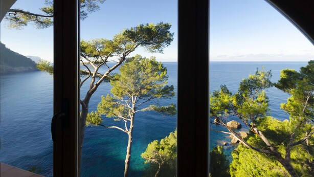 Une vue sur la côte, à travers une fenêtre, avec un ciel dégagé, une mer calme et des pins.