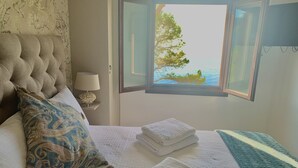 Une chambre à coucher avec une tête de lit capitonnée, un coussin à motifs, une lampe de chevet, une fenêtre donnant sur la mer et des serviettes soigneusement pliées.