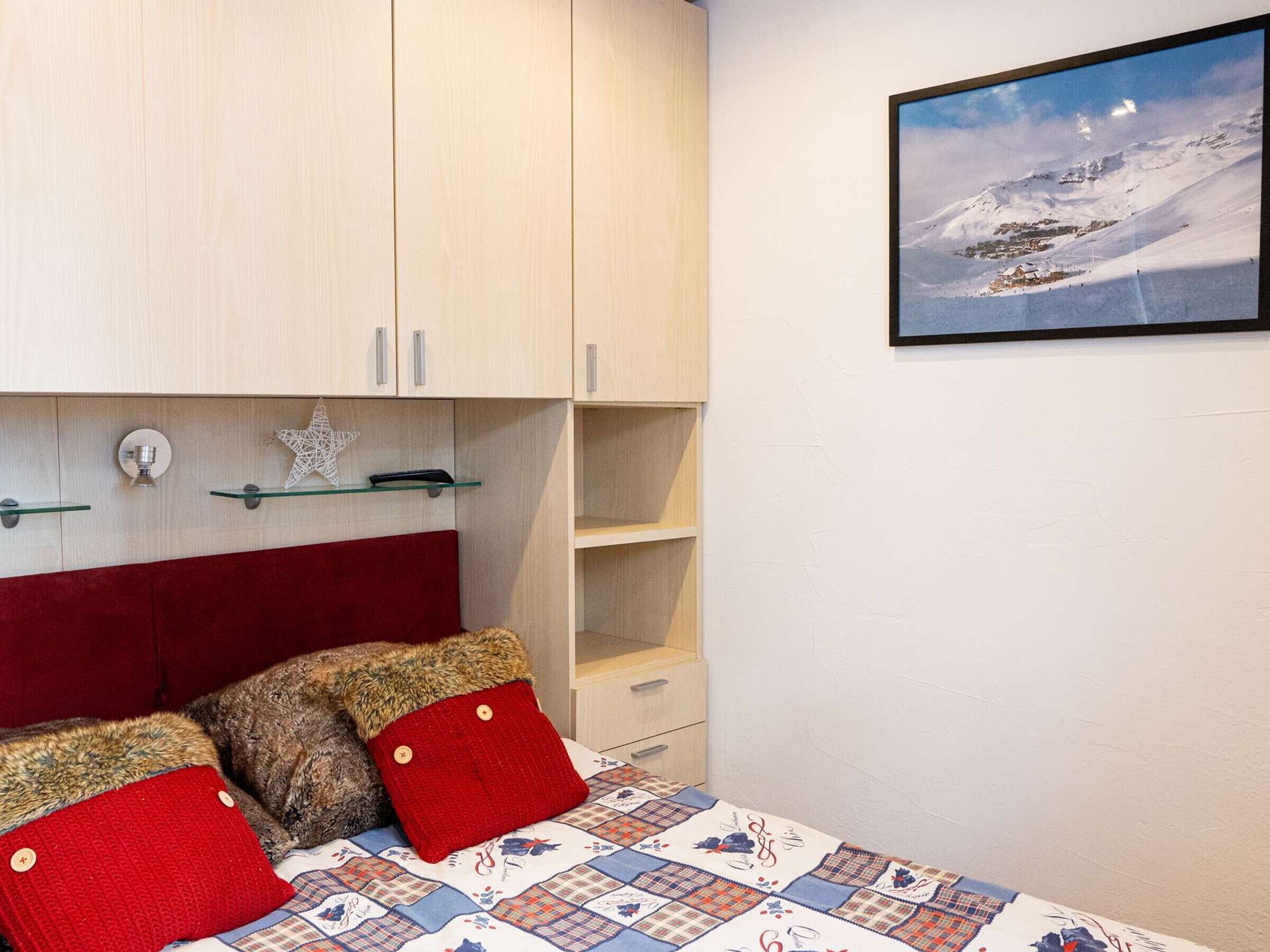 Un dormitorio con cama, cabecera roja, un cuadro colgado en la pared de una montaña nevada y un armario de madera con estantes de vidrio.