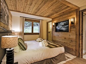 A cozy bedroom with a wooden ceiling, a bed with white linens, a mounted TV, and a window with curtains. - Luxury Chalet with 7 Bedrooms, Spa Access, and Stunning Mountain Views (Méribel)