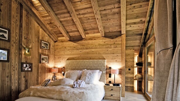 A cozy wooden room with a bed, a nightstand, and a fur-covered ottoman. - Luxury Chalet with 7 Bedrooms, Spa Access, and Stunning Mountain Views (Méribel)