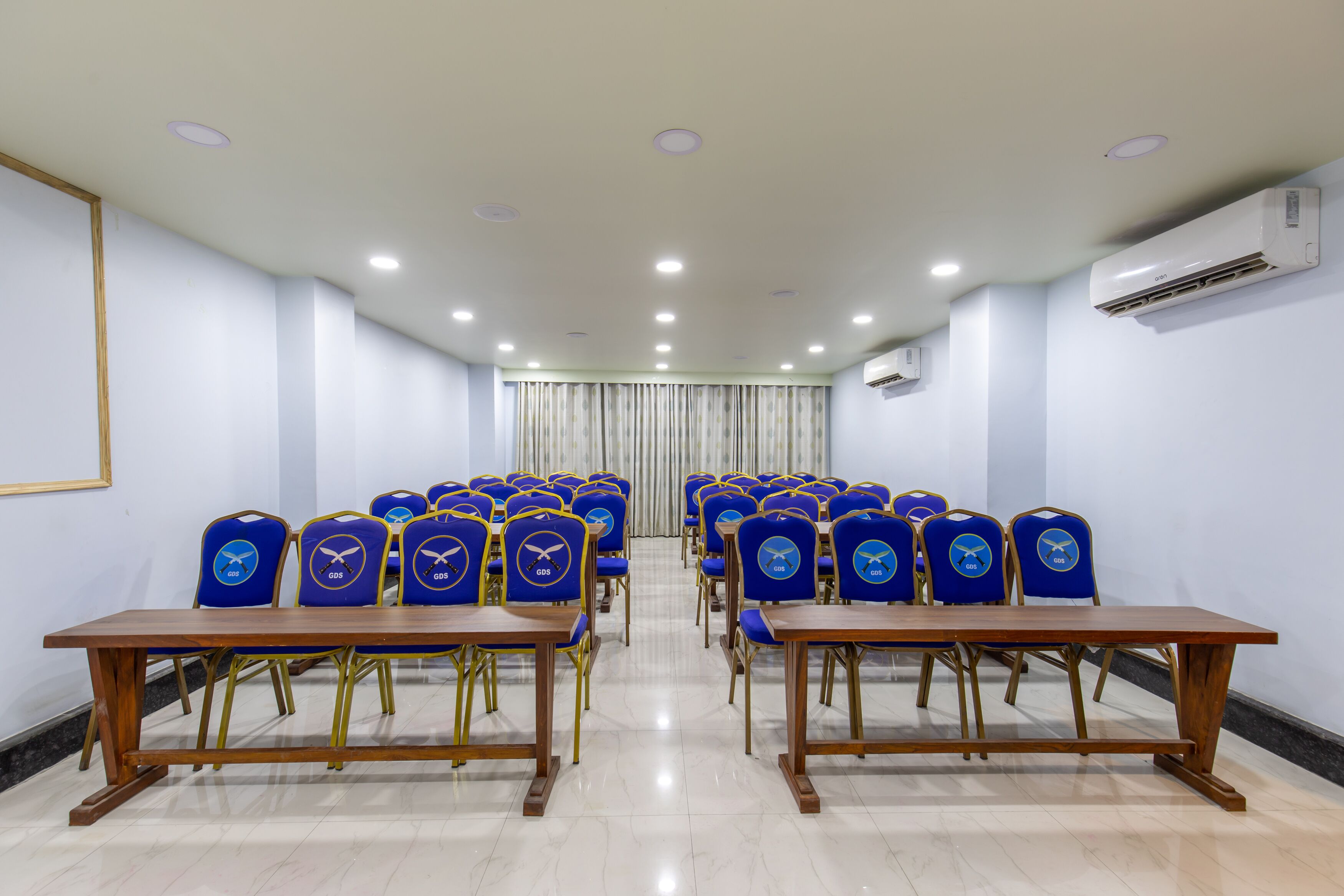 A conference room with rows of blue chairs and wooden tables, white walls, and ceiling lights.