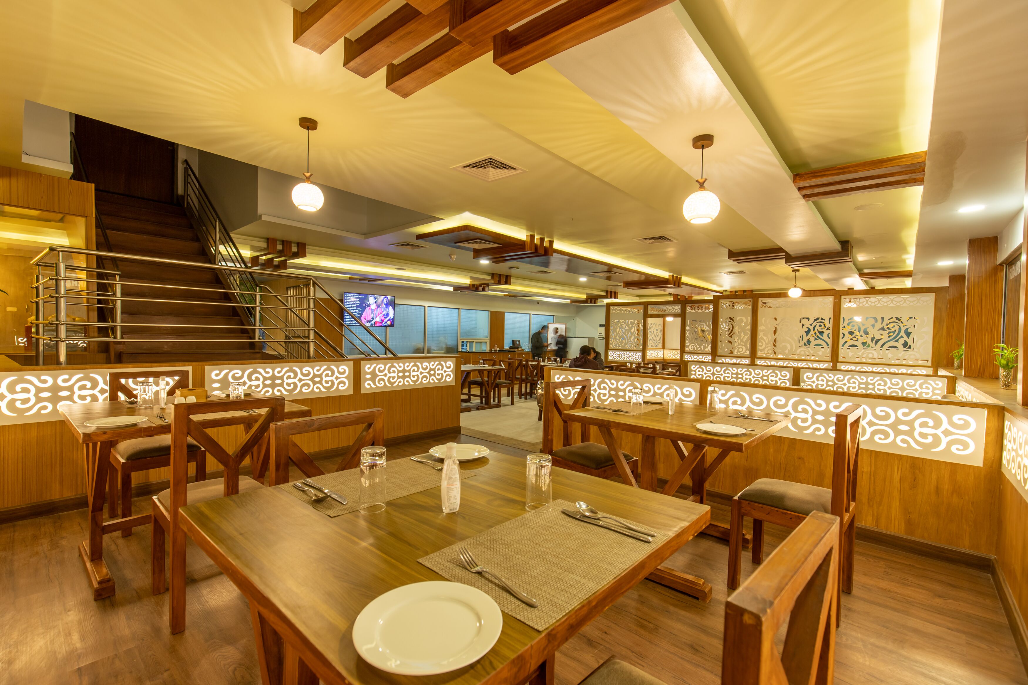 Restaurante com mobiliário de madeira, uma escada e painéis decorativos. Mesas montadas com pratos, talheres e copos.