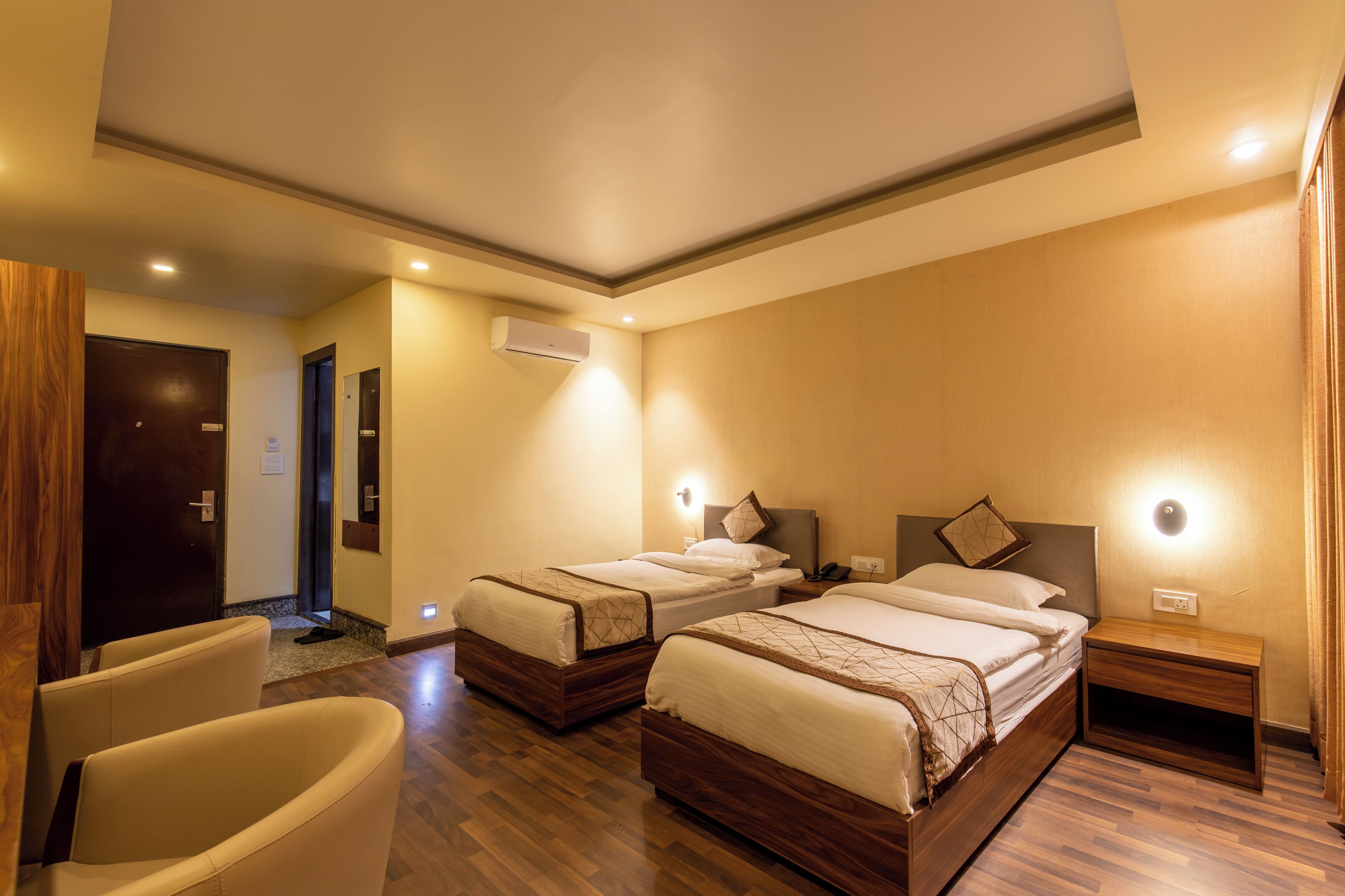 Habitación de hotel con dos camas, una zona de estar con una silla, piso de madera y una puerta que da a otra habitación.