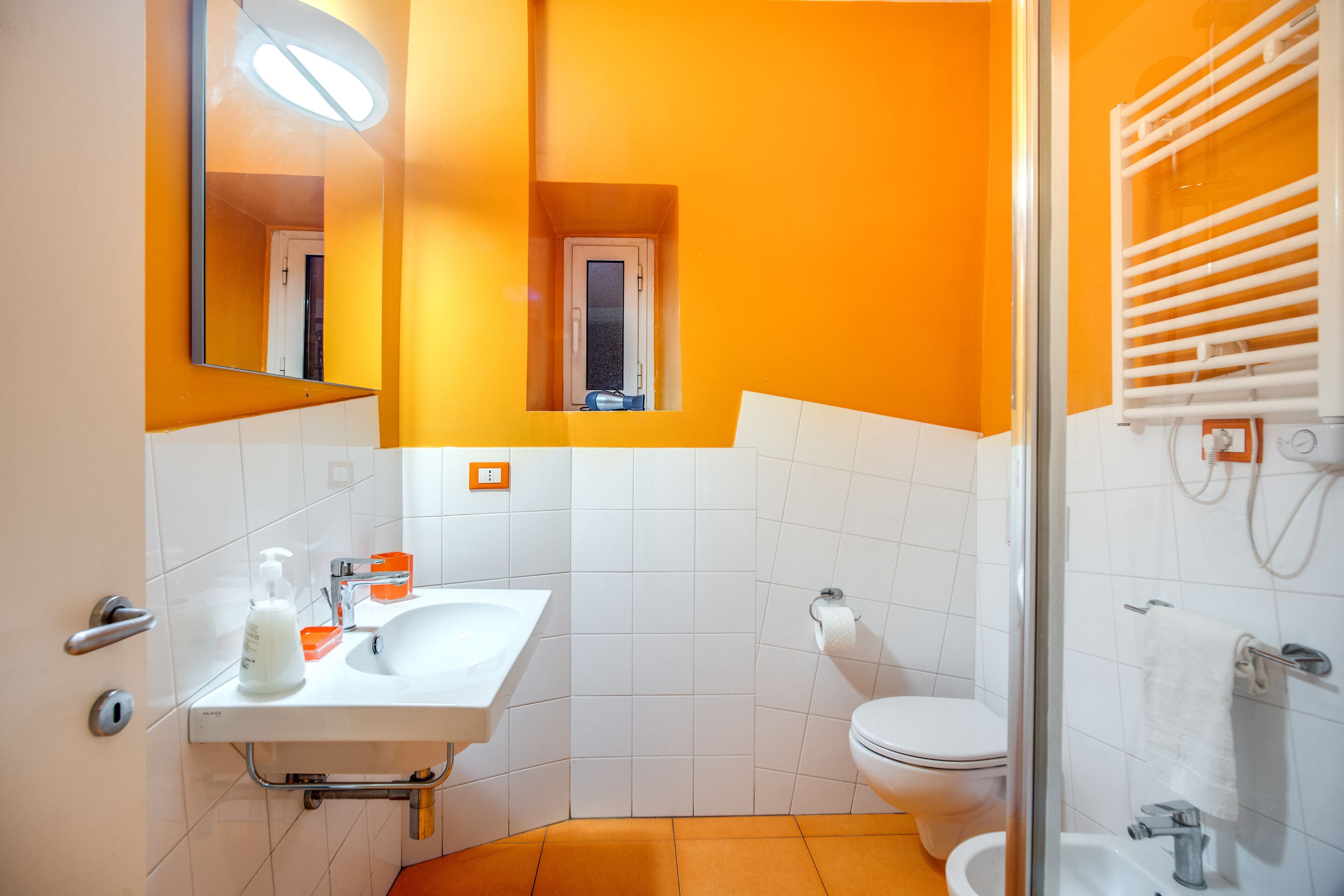 A bathroom with a white sink, mirror, and toilet, orange walls, and a glass shower enclosure.