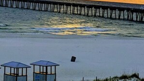 A beach with a pier extending into the water, two beach huts, and a sunset in the background. - Big Balcony beach front corner unit. Next to county pier. 4 Beach chair service! (Panama City Beach)