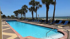 A rectangular swimming pool with blue water, surrounded by a tiled deck with a zigzag pattern. There are blue lounge chairs aligned along the pool's edge, and palm trees are visible in the background. - Big Balcony beach front corner unit. Next to county pier. 4 Beach chair service! (Panama City Beach)