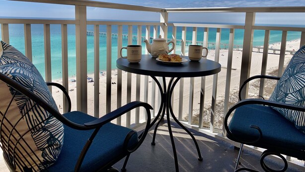 A balcony with a view of the beach, a table with coffee cups and a teapot, and two chairs with a tropical pattern. - Big Balcony beach front corner unit. Next to county pier. 4 Beach chair service! (Panama City Beach)